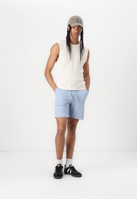 Letvægtscreme strikket ærmeløs sweater, lyseblå shorts, sorte sneakers med hvide detaljer og en grå kasket, som står mod en hvid baggrund.