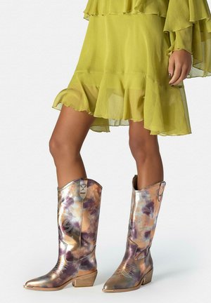 Mujer con botas de vaquero con patrón metálico y vestido amarillo con volantes, de pie contra un fondo liso.