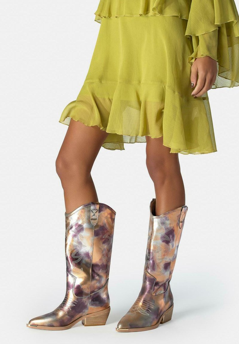 Frau trägt metallic gemusterte Cowboy-Stiefel und ein gelbes, gerafftes Kleid, sie steht vor einem schlichten Hintergrund.