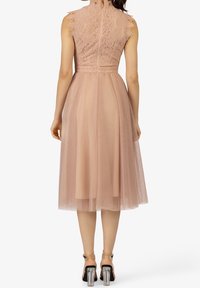 Femme portant une robe midi sans manches en dentelle rose et tulle avec une fermeture éclair au dos, associée à des sandales à talons blocs transparentes.