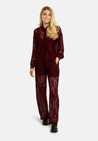 Bordový súprava z čipky obsahujúca košeľu na gombíky s dlhými rukávmi a nohavice so širokými nohavicami, zdobené kvetinovým vzorom a priehľadnou textúrou. Čierna obuv v cene.