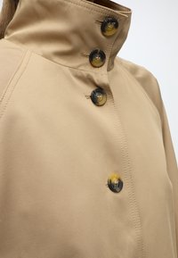 Gros plan d’un trench beige montrant un col montant avec quatre boutons ronds en écaille et des coutures détaillées sur un tissu lisse.