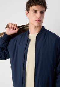Veste bomber zippée bleu marine avec une texture lisse, col côtelé et poches latérales, portée sur un pull en tricot beige, accompagnée d'un sac marron.
