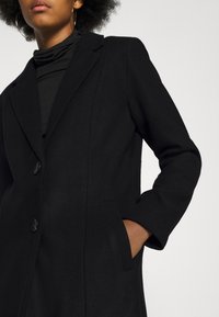 Manteau noir en mélange de laine avec une coupe ajustée, col cranté, deux boutons et poches latérales. Texture lisse avec une silhouette classique.