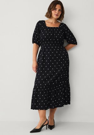 Kvinde iført en sort midi kjole med hvide prikker, firkantet halsudskæring, pufærmer og sorte slingback hæle, stående mod en ensfarvet væg.