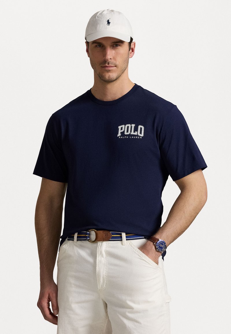 Tumši zili kokvilnas T-kreklis ar "POLO RALPH LAUREN" logo; kopā ar bežām šortiem un brūnu jostu. Redzams balts beisbola cepures un zilas pulksteņi.