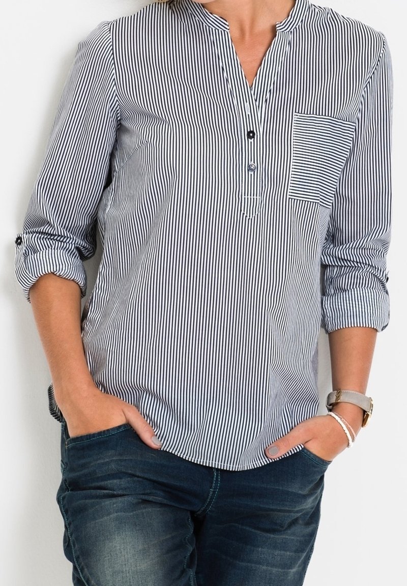 Camisa de rayas azul y blanca con cuello y tapeta de botones, bolsillo en el pecho y mangas enrolladas. Lleva puesta sobre unos jeans oscuros.