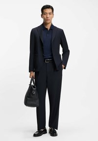 Granatowa marynarka i polo, dopasowane spodnie, czarne skórzane buty, trzymająca czarną torebkę o fakturze, minimalistyczny design, dopasowana sylwetka.