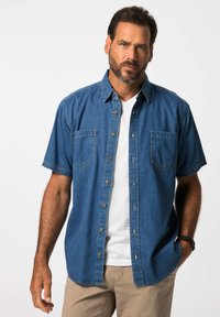 Korte mouwen denim shirt, donkerblauw met knopen aan de voorkant, twee borstzakken, gedragen met een wit T-shirt en beige broek.