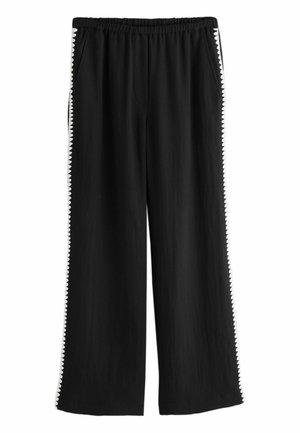 Mustat leveälahkeiset housut, joissa joustava vyötärö ja valkoinen siksakreunus molempien lahkeiden ulkosivuilla.