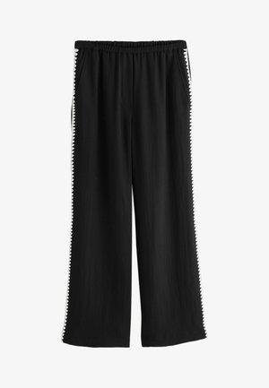 Mustat leveälahkeiset housut, joissa joustava vyötärö ja valkoinen siksakreunus molempien lahkeiden ulkosivuilla.