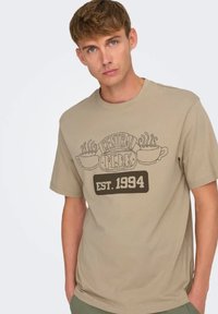 Camisa de algodón beige con un gráfico de "Central Perk" en marrón, que incluye un diseño de tazas y vapor. Manga corta, corte relajado, cuello redondo.