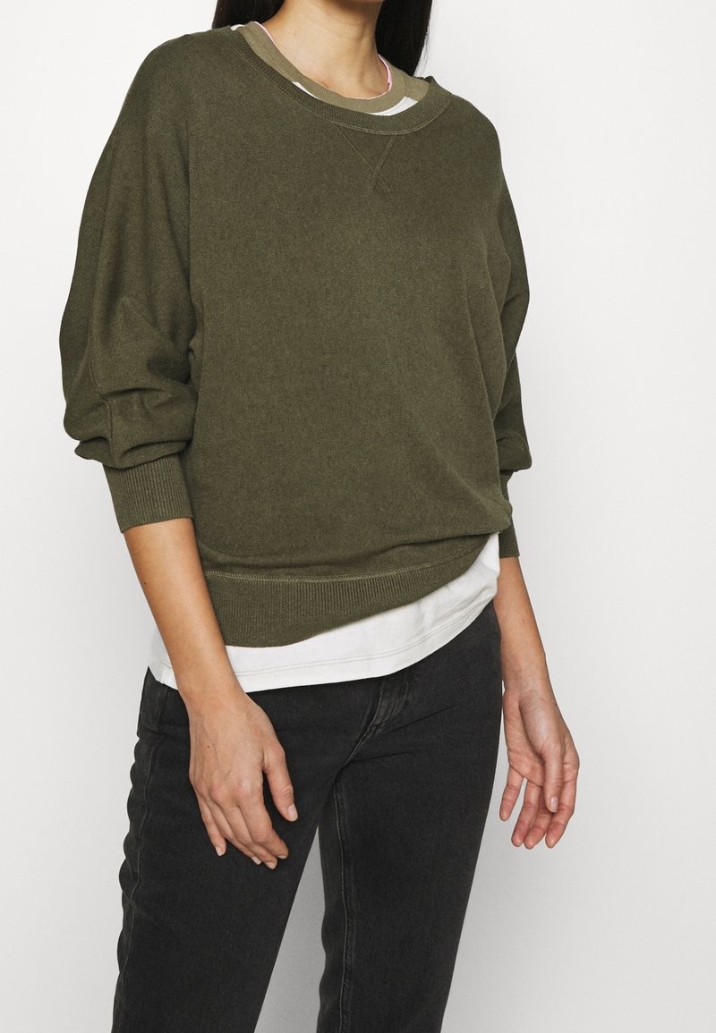 Olijfgroen pullover trui met een ontspannen pasvorm, geribbelde mouwen en zoom, gedragen over een witte shirt, toont een zachte textuur en subtiel ontwerp.