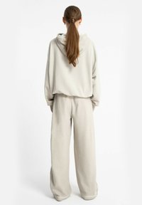 Kobieta z długimi brązowymi włosami związanymi w kok, ubrana w luźny beżowy hoodie i szerokie spodnie, stojąca przodem do białego tła.