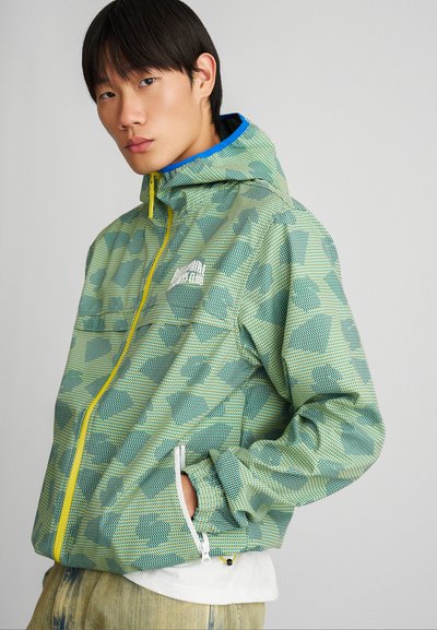 Chaqueta ligera con un patrón verde que incluye capucha, cremallera amarilla y bolsillos laterales. Material texturizado con un acabado similar a una malla.