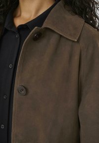 Brown suede jacket with a collar, featuring a button closure and visible stitching details. Worn over a dark shirt.