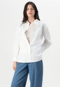 Bela srajca z gumbki iz bombaža, ohlapnega kroja, z enim žepom na prsih in dolgimi rokavi, v kombinaciji s svetlo modrimi kavbojkami.