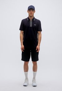 Μαύρο πόλο μπλουζάκι με λευκές λεπτομέρειες, συνδυασμένο με μαύρες βερμούδες. Το μοντέλο φοράει καπέλο και λευκά αθλητικά παπούτσια με εμφανείς ριμπ κάλτσες. Απλός σχεδιασμός.