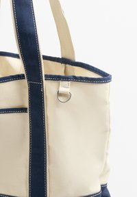 Platnena torba z mornarsko modrimi akcenti, ki ima trdne ročaje, vidne šive in kovinski obroč za pritrditev. Kremaste barve.