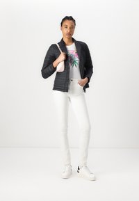 Casaco preto acolchoado sobre uma t-shirt branca com design floral, combinado com jeans brancos e ténis com detalhes em preto. Detalhe da bolsa rosa claro.