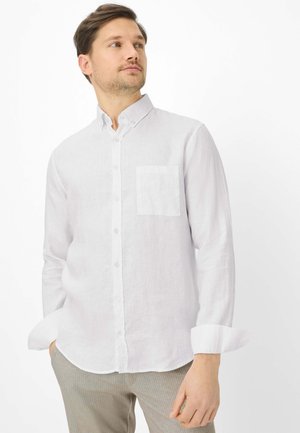 Uomo che indossa una camicia bianca a maniche lunghe con taschino sul petto, pantaloni chiari, mani in tasca, guarda di lato.