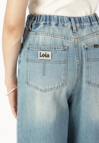 Osoba v světlomodrých džínech s elastickým pasem a nášivkami loga "Lois" na zadních kapsách, bílém topu a perlouvém náramku na zápěstí.