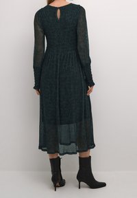 Lång, mörk teal klänning med smockad bodice och genomskinliga långa ärmar. Har en nyckelhålsdetalj bak och en flödande, texturerad kjol. Bärs med svarta klackstövlar.