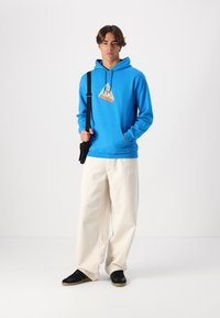 Blue hoodie with a triangular graphic design, paired with off-white wide-leg pants and black shoes. The fabric appears soft and casual.