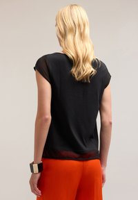 Zwarte blouse met doorschijnende korte mouwen, ronde halslijn en een iets losse pasvorm. Gepaard met oranje brede broek en een geometrische armband.
