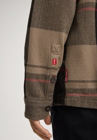 Brązowa koszula kurtka w jodełkę w kratę z czerwonymi akcentami, zapinanymi mankietami oraz naszywką na rękawie z czerwonym wykrzyknikiem.