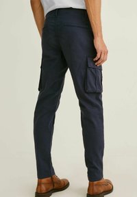 Man klädd i marinblå cargobyxor med fällfickor, bruna läderstövlar, stående mot en enkel ljus bakgrund, sedd bakifrån.