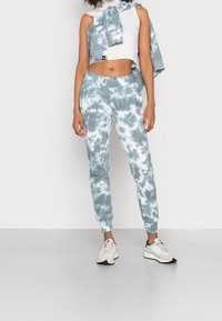 Světle modré tie-dye tepláky s elastickými manžetami, v kombinaci s bílým žebrovaným crop topem a odpovídajícím tie-dye svetrem přehozeným přes ramena.