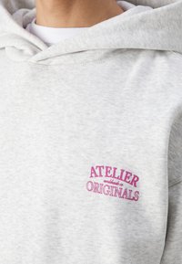Sweat à capuche gris clair avec texte rose "Atelier Originals" brodé sur la poitrine, porté sur une chemise blanche.