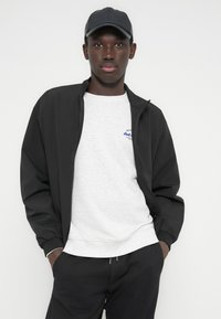 Μαύρο μπουφάν bomber με φερμουάρ, φορεμένο πάνω από ένα ανοιχτό γκρι φούτερ. Διαθέτει μικρό μπλε λογότυπο. Συνδυάζεται με σκούρο παντελόνι και μαύρο καπέλο.