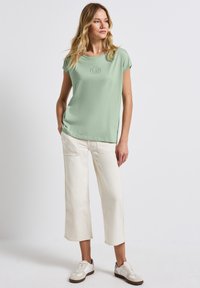 Mátově zelené tričko s krátkým rukávem s jemným nápisem "PLAIN", kombinované s krémovými zkrácenými kalhotami a bílými teniskami s hnědými detaily.