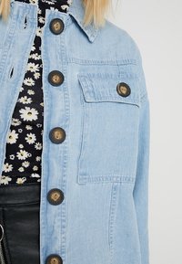 Světle modrá džínová bunda s klasickým límcem, zapínáním na knoflíky, jednou náprsní kapsou a volnějším střihem. Vzoru látky viditelné pod ní.