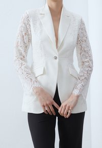 Biała marynarka z koronkowymi rękawami, dopasowany krój, zapięcie na jeden guzik i dwie przednie kieszenie. Noszona na czarnych spodniach.