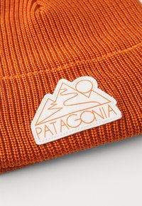 Narancssárga kötött sapka bordázott textúrával. Fehér címkével rendelkezik, amelyen hegydesign és narancsszínű "PATAGONIA" szöveg látható.