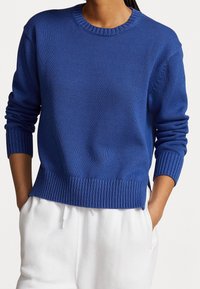 Personne portant un pull en laine bleu avec des poignets et un ourlet côtelés, associé à un pantalon blanc à cordon de serrage, les mains dans les poches.