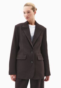 Blazer marrón a medida hecho de tejido texturizado, con solapa de muesca, dos bolsillos frontales y dos botones negros.