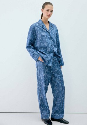 Jeune femme aux cheveux attachés, portant un haut et un pantalon de pyjama bleu à motifs floraux assortis, mains dans les poches, debout contre un mur clair uni.
