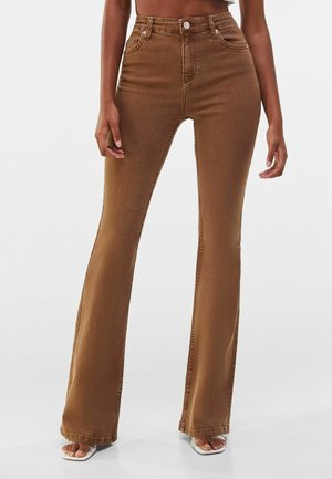 Kobieta ubrana w brązowe dzwony z wysokim stanem i białe sandały z odkrytymi palcami, stojąca na jednolitym tle.