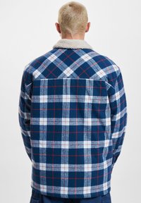 Rutet flannel jakke i navy og hvid med røde detaljer, der har en sherpa-krave og en afslappet pasform, vist fra bagsiden.
