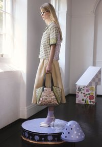 Kobieta w okularach i kwiecistej koszuli oraz beżowej spódnicy, trzymająca dopasowaną kwiecistą torebkę, stoi na pomalowanej na fioletowo platformie wewnątrz budynku.