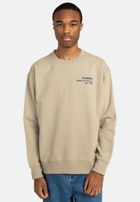 Sudadera beige con cuello redondo, corte oversize, puños y dobladillo de canalé. Presenta un logo en texto azul oscuro en el pecho.