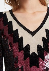 Haut à col en V orné de sequins avec un motif en zigzag noir, bordeaux et beige ; tissu doux avec des sequins texturés et brillants de différentes couleurs.