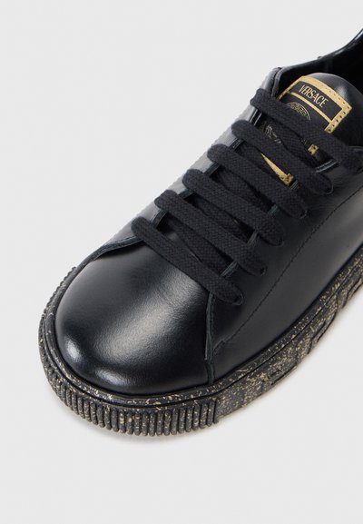 Zapatilla de cuero negro con punta redonda, suela gruesa texturizada con motas doradas y cordones planos negros. Logo dorado en la lengüeta.