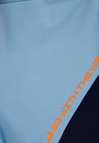 Svetlomodrá tkanina s hladkým povrchom a tmavomodrým akcentom. Obsahuje oranžové heslo "CHOĎ S PRÚDOM" pozdĺž švu.