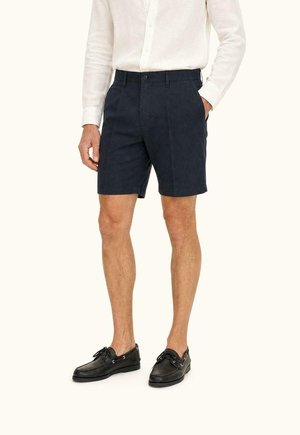 Man draagt een wit overhemd met lange mouwen in donkerblauwe shorts met de handen in de zakken, gecombineerd met zwarte bootschoenen, staat rechtop.