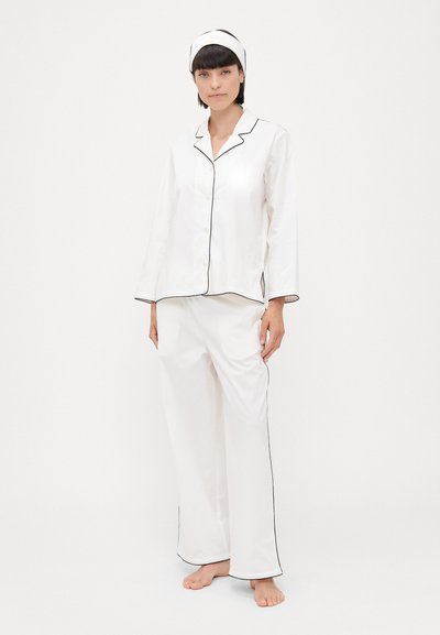 Set de pijama alb, compus dintr-o bluză cu mâneci lungi și guler închis cu nasturi și pantaloni largi, accentuat cu piping negru pe margini. Material moale cu o textură netedă.